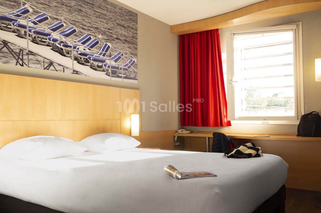 Chambre d'hôtel lumineuse avec un lit double, une fenêtre avec rideaux rouges et un bureau en bois clair.