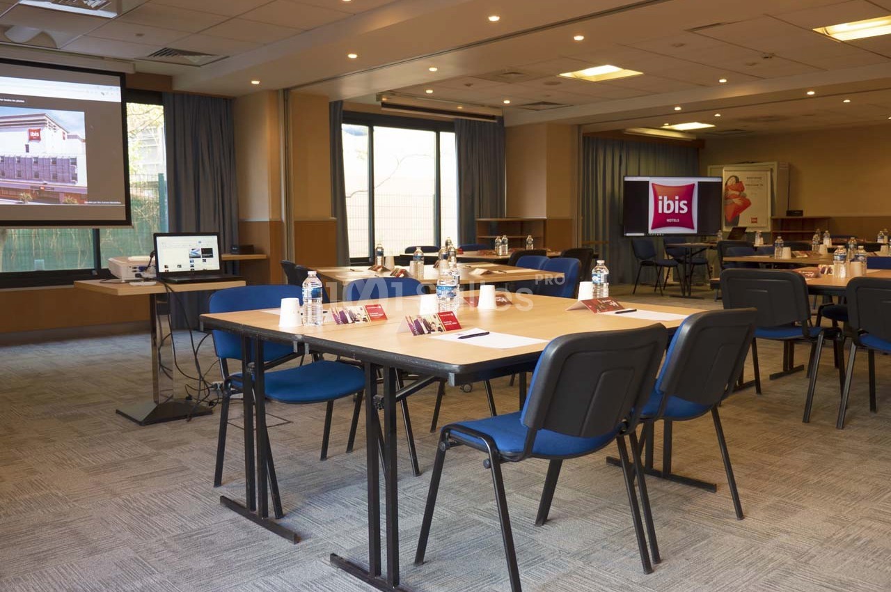 Salle de réunion lumineuse avec tables disposées en U, chaises bleues, écran de projection et matériel de présentation.