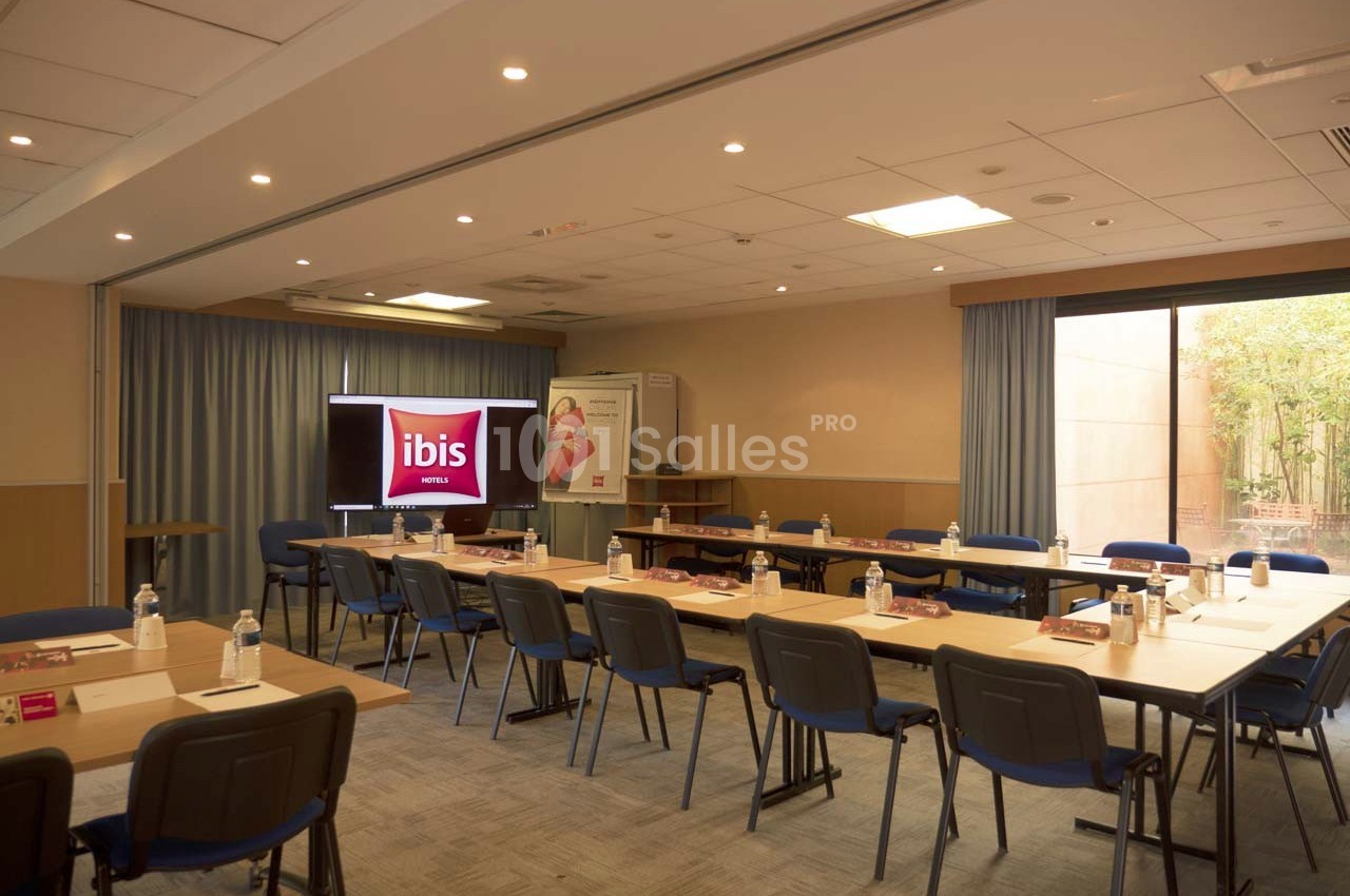 Salle de réunion lumineuse avec tables disposées en U, chaises, écran de présentation et bouteilles d'eau.