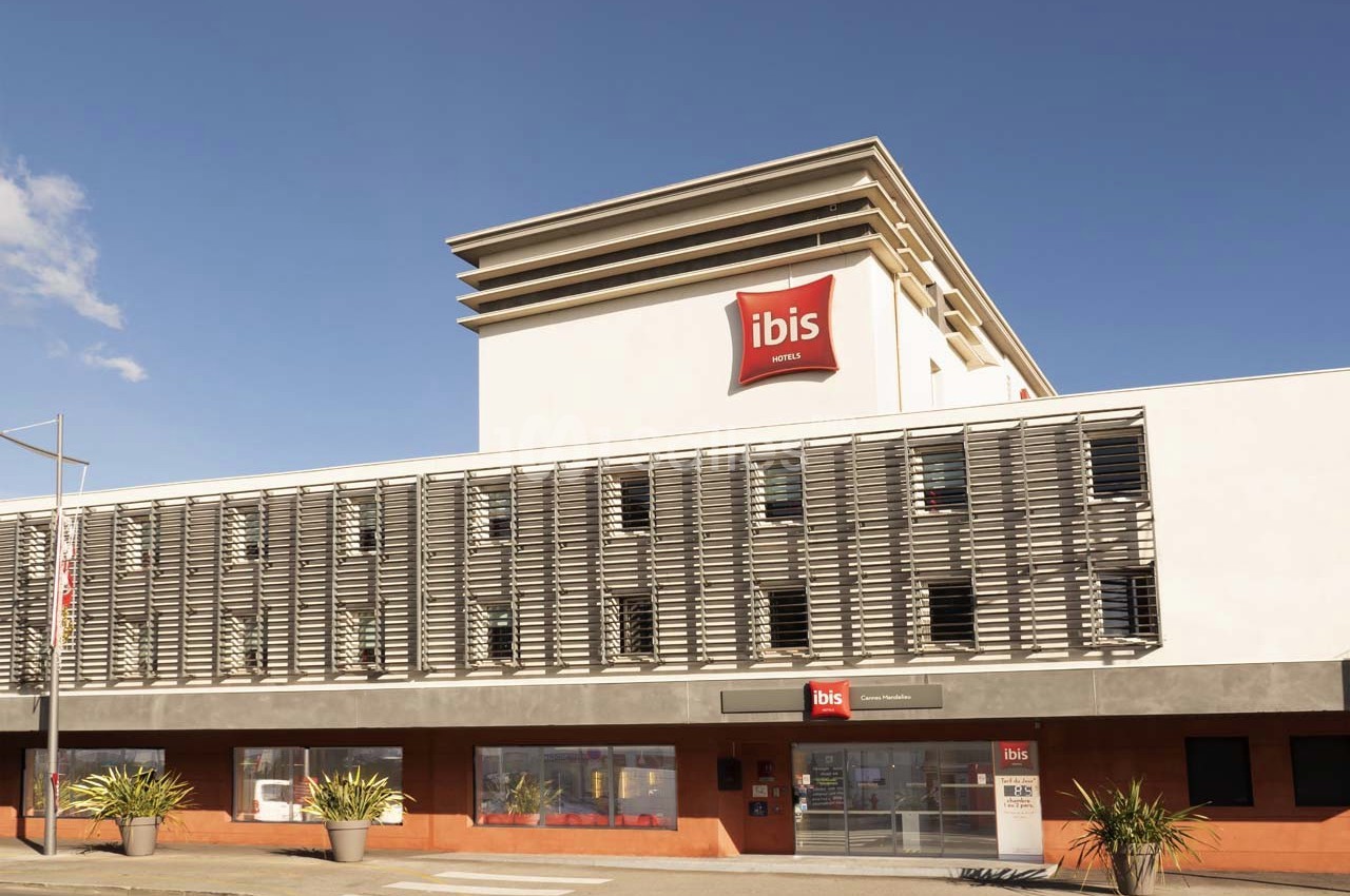 Façade d'un hôtel Ibis moderne avec des volets métalliques et un logo rouge visible sur le toit.