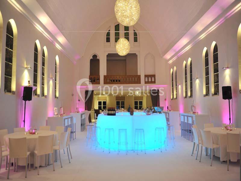 Salle lumineuse avec un bar central éclairé en bleu, entouré de tables et chaises blanches, dans un décor moderne.