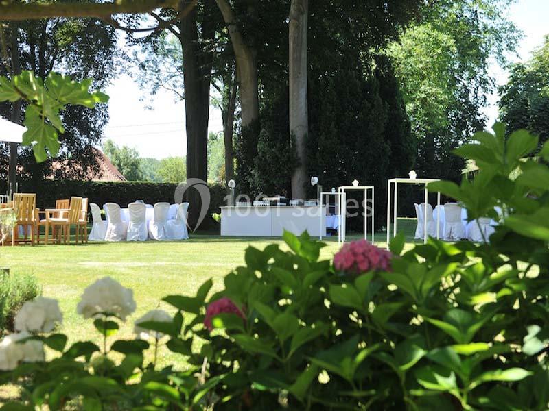 Jardin verdoyant avec tables et chaises blanches disposées pour un événement en plein air, entouré de végétation.