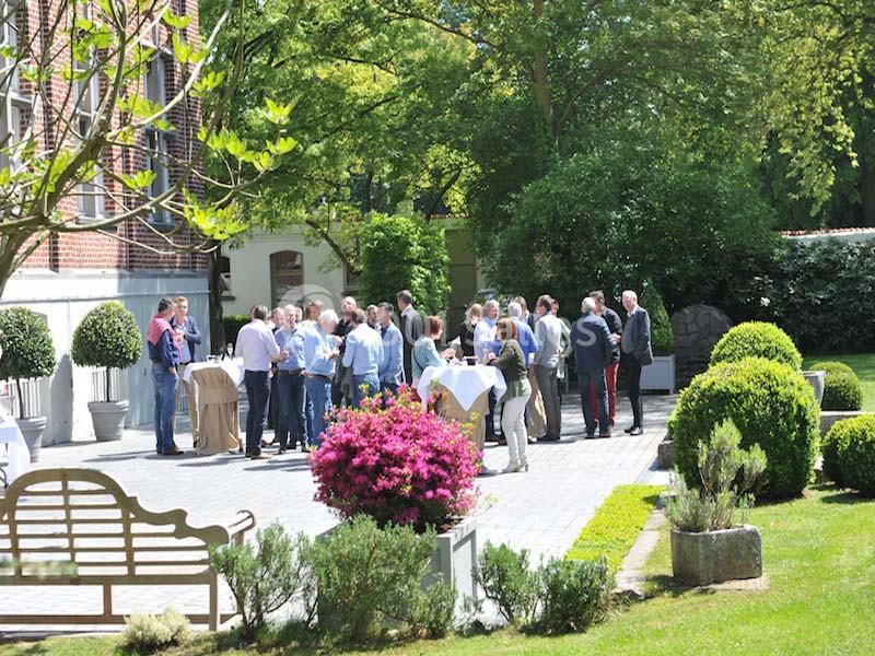 Groupe de personnes discutant debout autour de tables dans un jardin avec des arbustes et des arbres en arrière-plan.