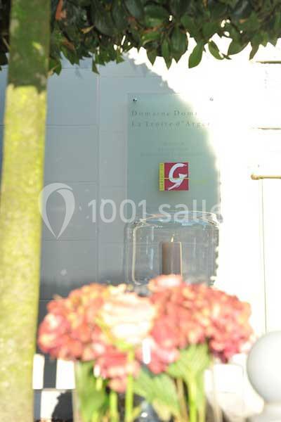 Plaque murale avec un logo rouge et jaune, entourée de végétation et de fleurs floues au premier plan.
