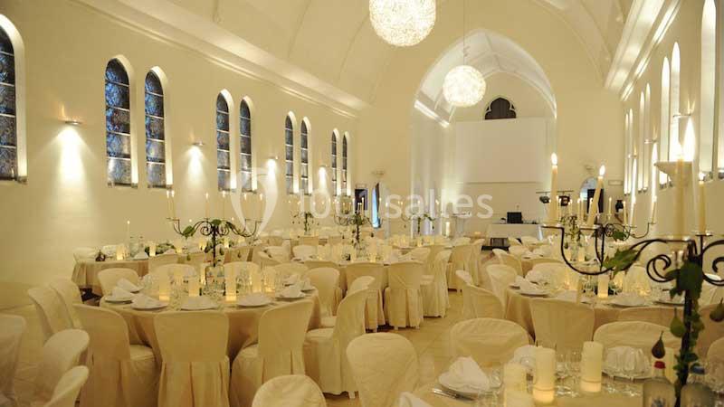 Salle de réception élégante avec tables rondes décorées de nappes blanches, chandeliers et éclairage chaleureux.