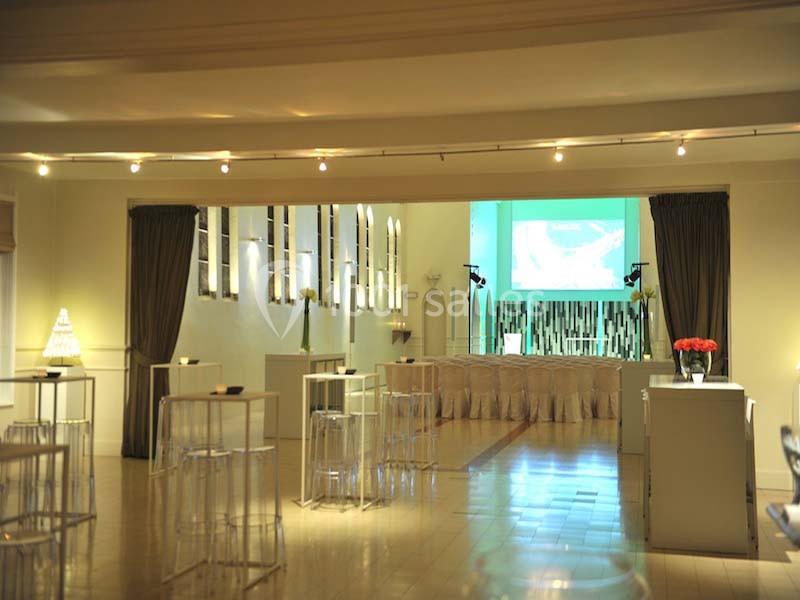 Salle lumineuse aménagée pour un événement, avec tables hautes, rideaux sombres et écran de projection en fond.