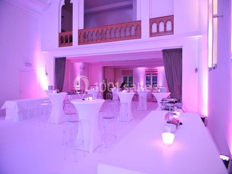 Salle lumineuse avec tables hautes, chaises transparentes et éclairage rose, décorée pour un événement.