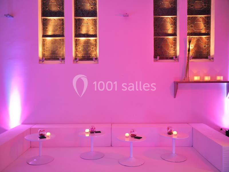 Banquettes blanches avec petites tables rondes, éclairage rose et bougies, devant des vitraux illuminés.
