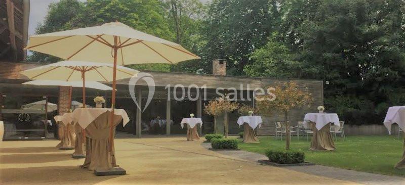 Location salle Brunehaut (Hainaut) - Le Clos de la Conciergerie #5 Terrasse aménagée avec des tables hautes couvertes de nappes blanches et des parasols, entourée de verdure.
