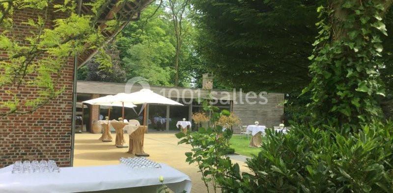 Location salle Brunehaut (Hainaut) - Le Clos de la Conciergerie #3 Espace extérieur avec tables dressées sous des parasols, entouré de verdure et d'un bâtiment en bois.