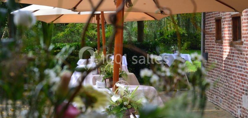 Location salle Brunehaut (Hainaut) - Le Clos de la Conciergerie #11 Terrasse extérieure avec tables dressées sous des parasols, entourée de verdure et d'un mur en briques rouges.