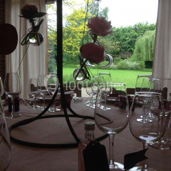 Location salle Brunehaut (Hainaut) - Le Clos de la Conciergerie #10 Table décorée avec des verres, des fleurs suspendues et vue sur un jardin verdoyant à travers de grandes fenêtres.