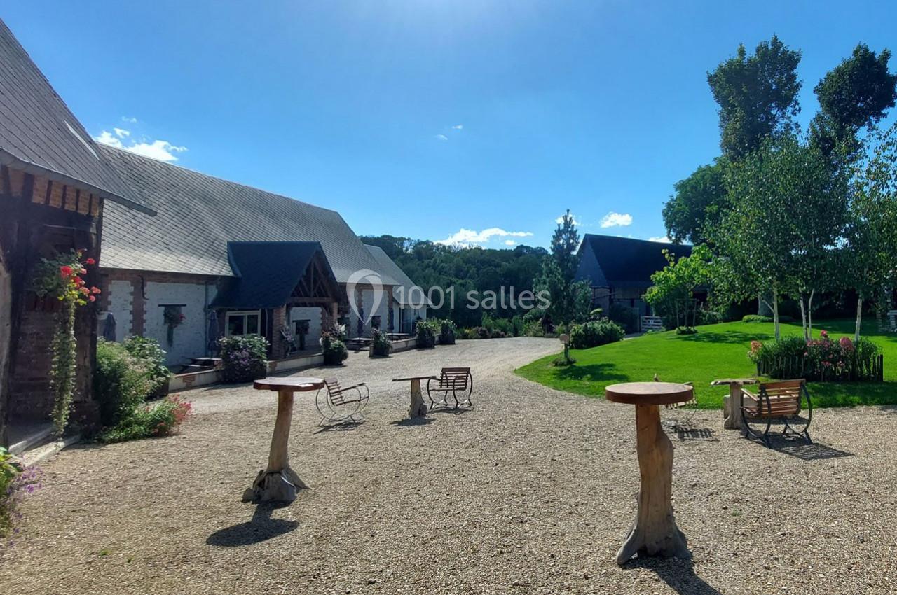 Cour extérieure avec tables en bois, chaises et bâtiments en pierre, entourée de verdure sous un ciel bleu.