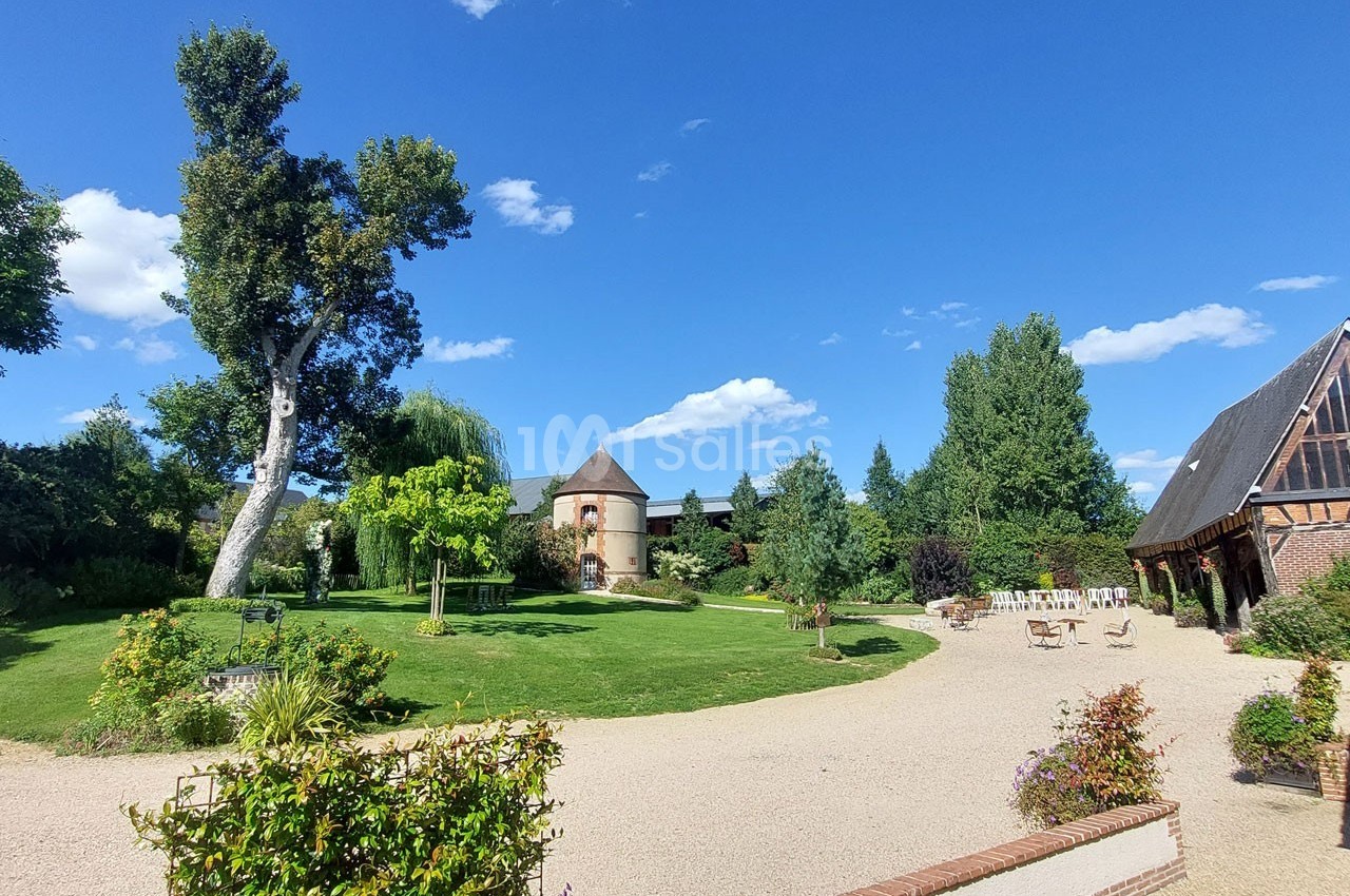 Jardin verdoyant avec pelouse, arbres, allée gravillonnée, bâtiment en briques et tour ronde sous un ciel bleu.