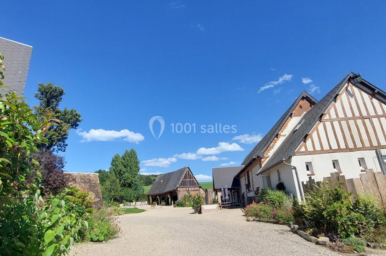 Cour d'une ferme normande avec bâtiments à colombages, jardin fleuri et ciel bleu dégagé en arrière-plan.