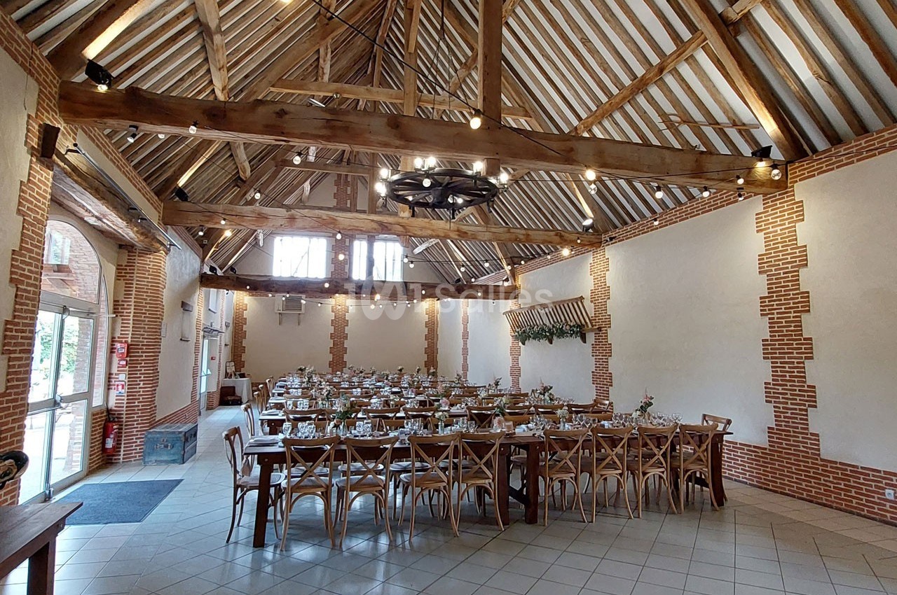 Salle de réception avec charpente en bois apparente, tables dressées et chaises en bois, éclairée par des fenêtres et des…