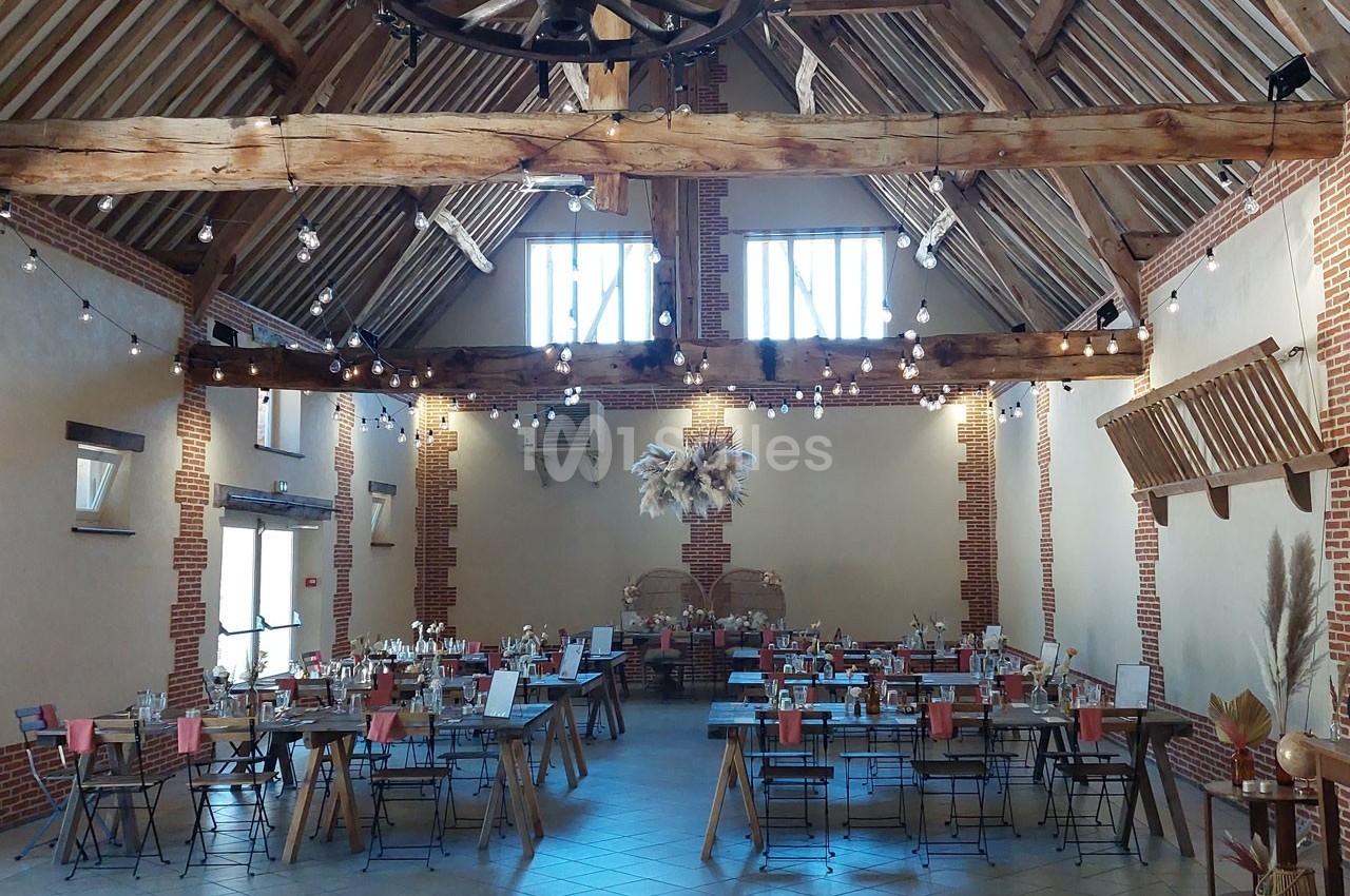 Salle de réception avec poutres apparentes, tables dressées et décorations suspendues dans un cadre rustique.
