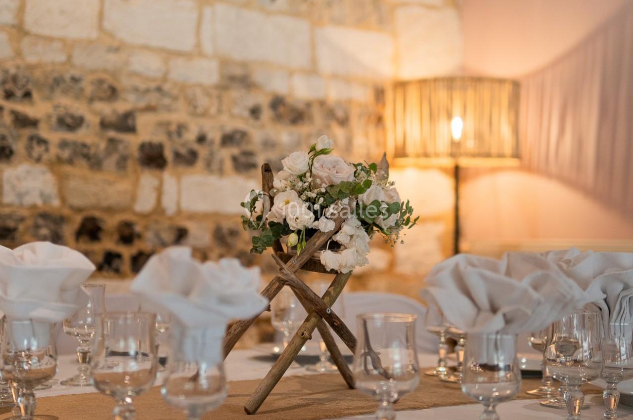 Centre de table floral avec roses blanches et feuillage, posé sur une table dressée dans une salle en pierre éclairée.