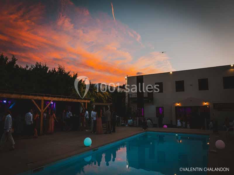 Soirée en extérieur au bord d'une piscine éclairée, avec invités sous un ciel coloré au coucher du soleil.