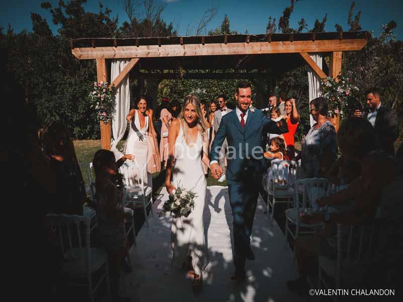 Un couple souriant marche sous une pergola en bois lors d'une cérémonie de mariage en extérieur, entouré d'invités.