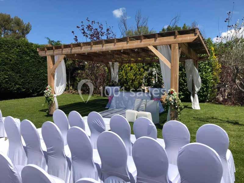 Pergola en bois décorée pour une cérémonie en plein air, entourée de chaises blanches et de fleurs.