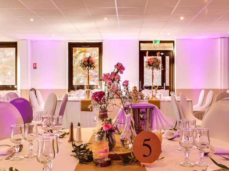 Salle de réception décorée avec des tables dressées, des fleurs roses et une lumière d'ambiance violette.