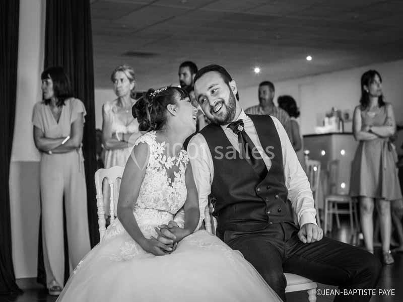 Un couple assis, souriant et complice, entouré d'invités debout dans une salle de réception.