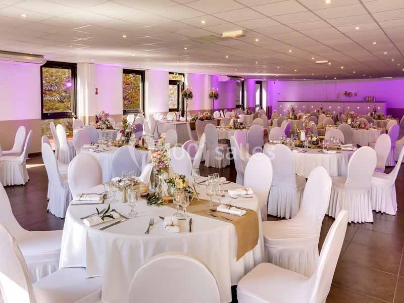 Salle de réception décorée avec des tables rondes, nappes blanches, chaises houssées et éclairage violet.