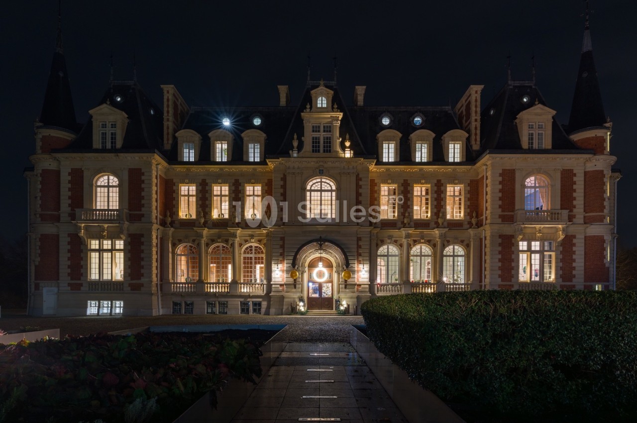 Façade d'un château illuminé de nuit, avec des fenêtres éclairées et un chemin menant à l'entrée principale.