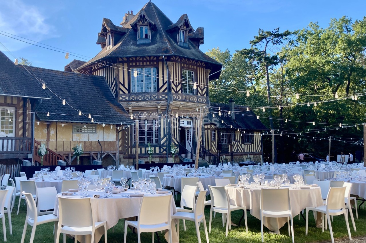 Tables dressées pour un événement en extérieur devant une grande maison à colombages, sous des guirlandes lumineuses.