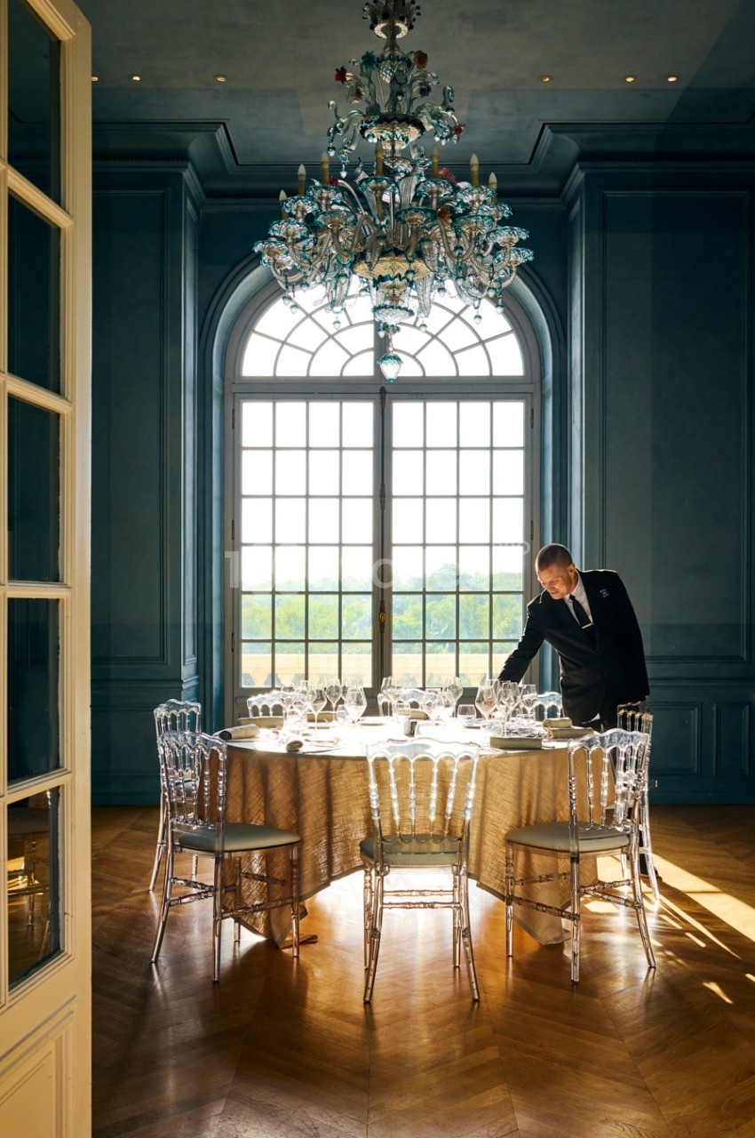 Un serveur ajuste une table élégamment dressée dans une salle lumineuse avec un grand lustre et des fenêtres cintrées.