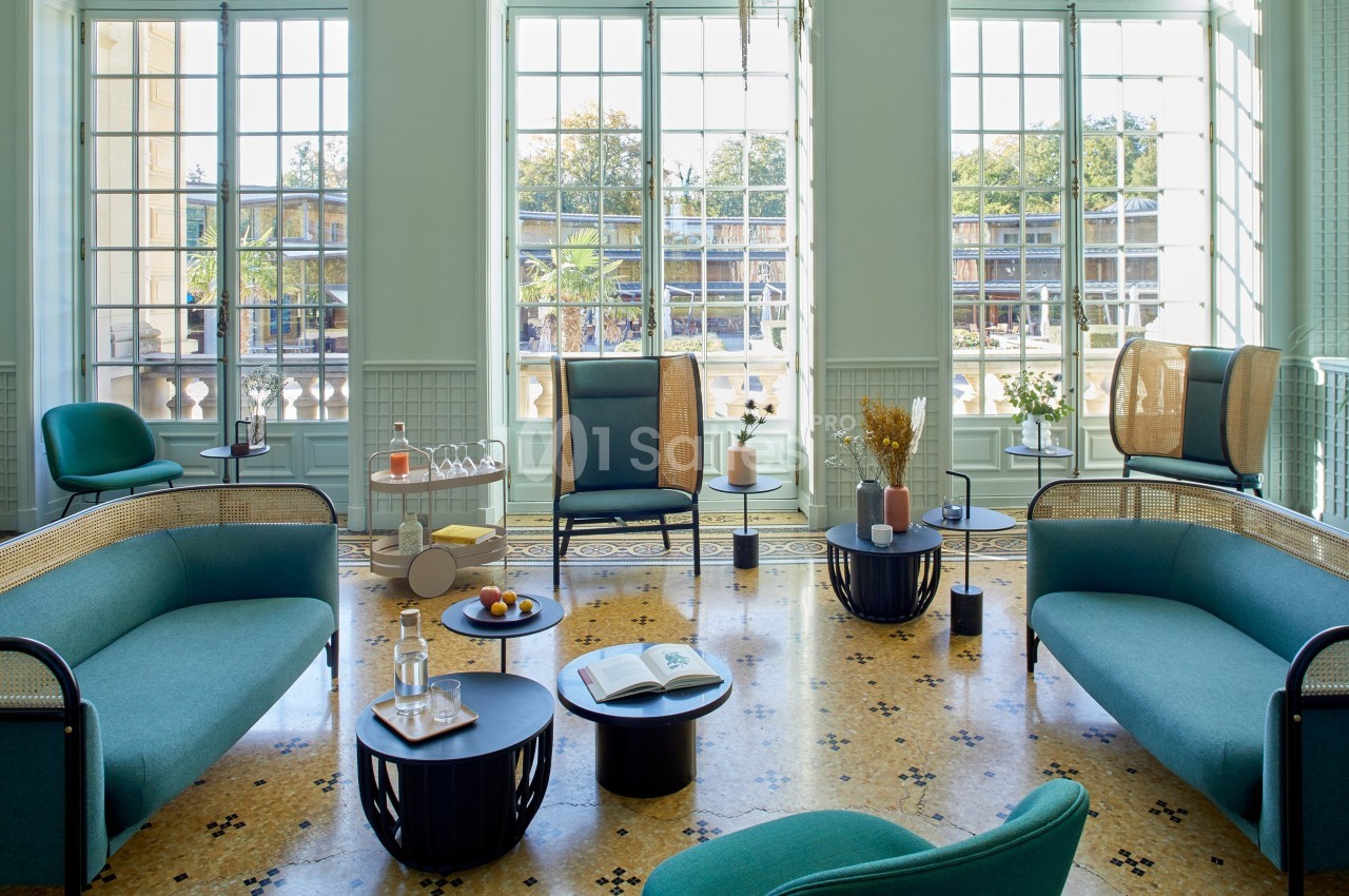 Salon lumineux avec mobilier moderne, canapés verts, tables basses et grandes fenêtres donnant sur un extérieur arboré.