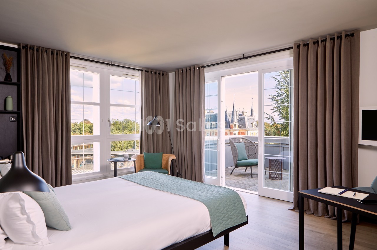 Chambre lumineuse avec lit double, bureau, fauteuil, grandes fenêtres et balcon offrant une vue sur un château.