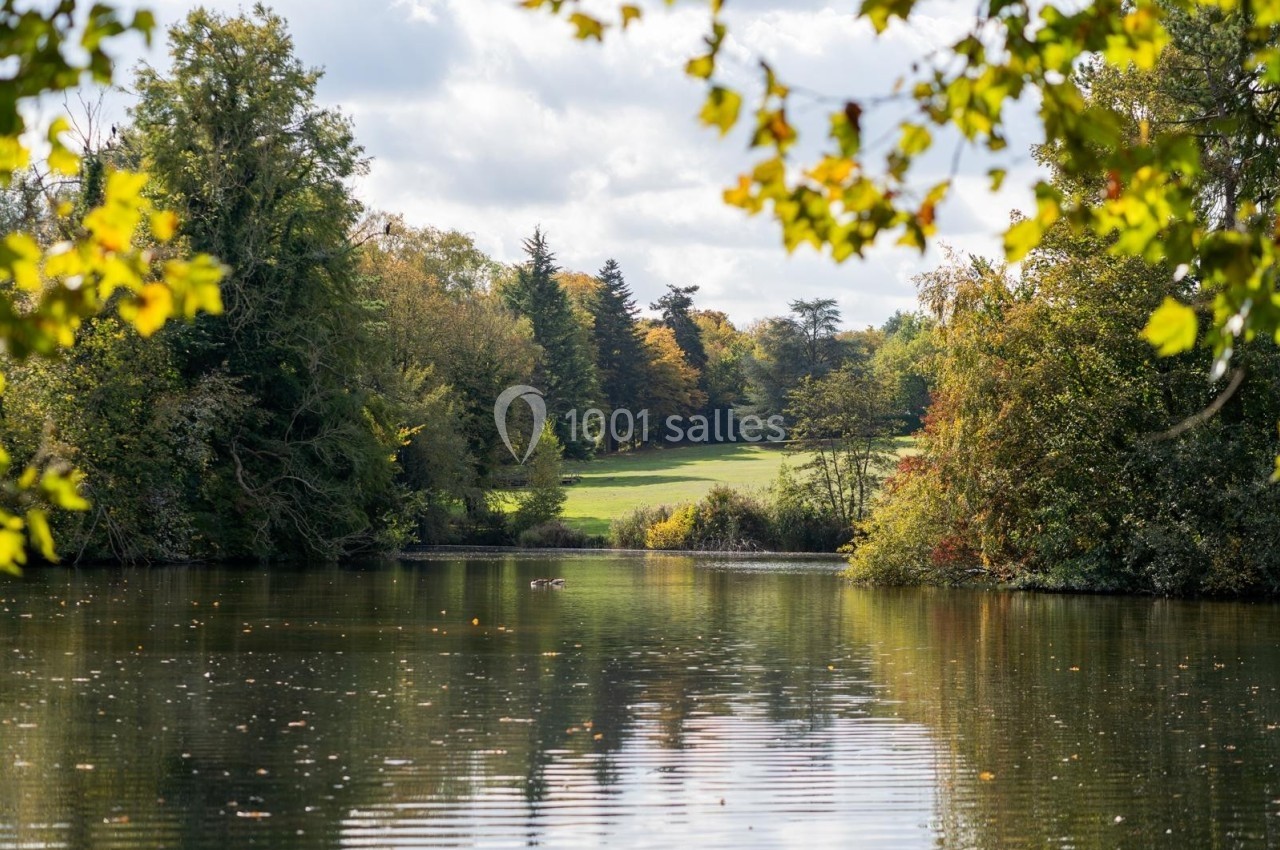 Vue d'un lac entouré d'arbres aux feuillages d'automne, avec une prairie en arrière-plan sous un ciel partiellement nuageux.