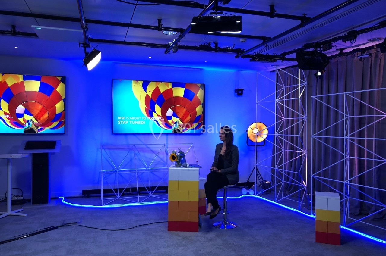 Une femme assise dans un studio coloré avec des écrans affichant une montgolfière et des éclairages lumineux.
