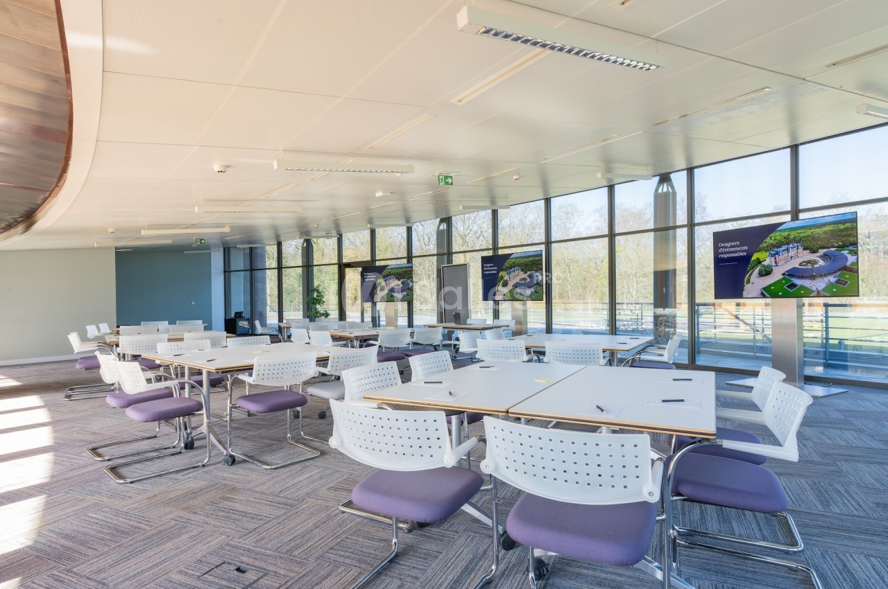 Salle de réunion lumineuse avec des tables blanches, des chaises violettes et des écrans affichant des présentations.