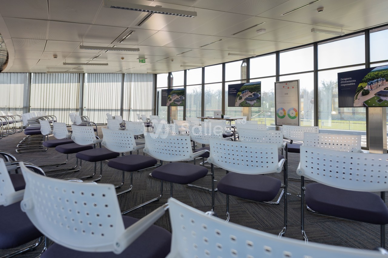 Salle de conférence lumineuse avec des rangées de chaises blanches et des écrans affichant des présentations.