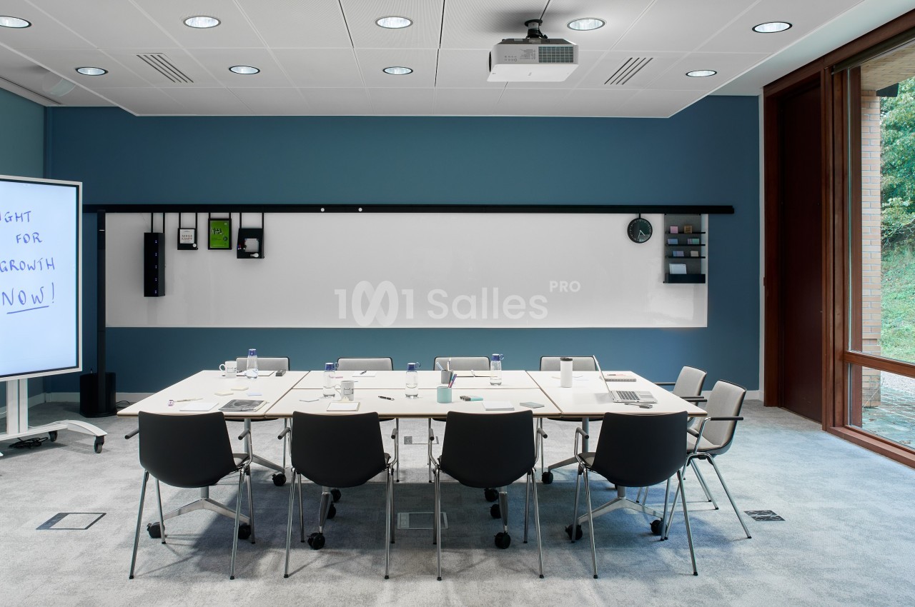 Salle de réunion moderne avec table, chaises, tableau blanc, écran interactif et vue sur un espace vert extérieur.