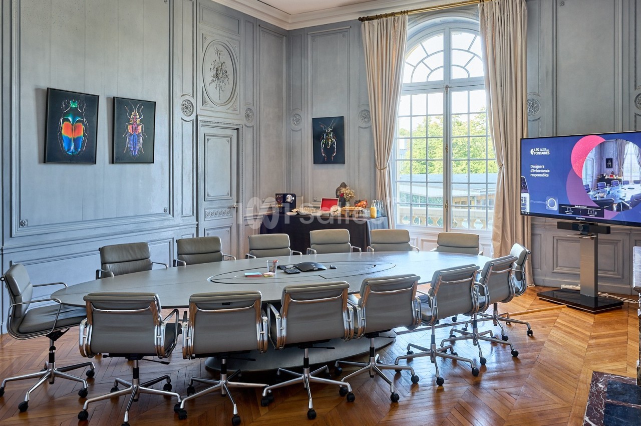 Salle de réunion élégante avec grande table ovale, chaises modernes, écran de présentation et fenêtres lumineuses.