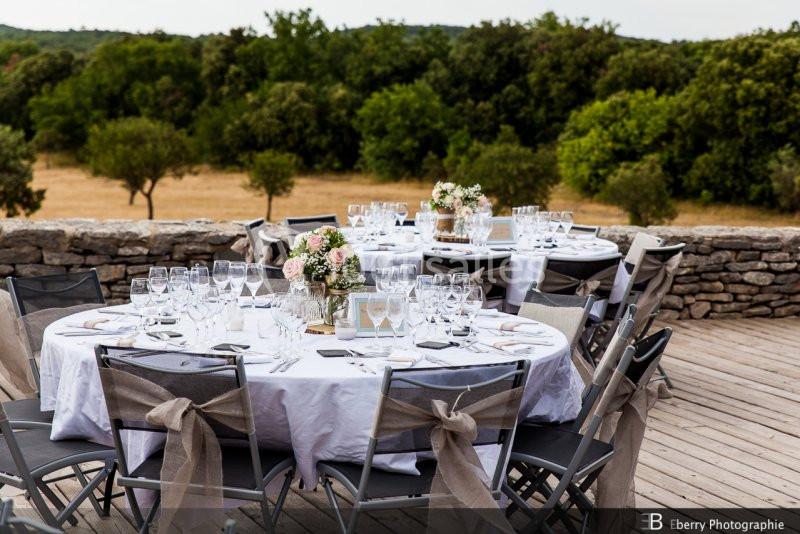 Table ronde dressée pour un événement en extérieur, avec nappes blanches, verres, fleurs et chaises décorées.