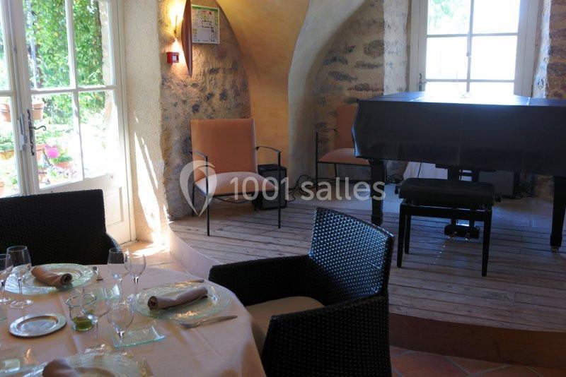 Salle lumineuse avec un piano noir sur une estrade, des chaises et une table dressée près d'une fenêtre.
