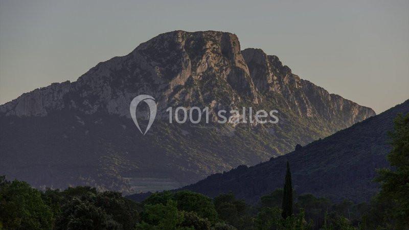 Montagne rocheuse éclairée par une lumière douce, entourée de collines boisées et d'arbres au premier plan.