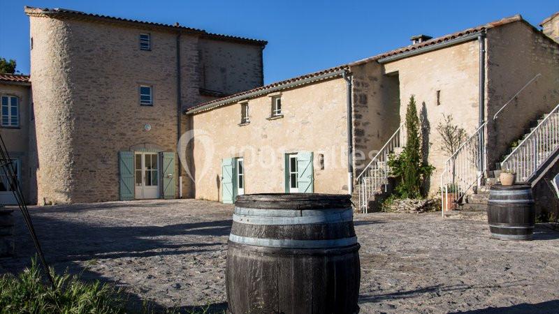 Cour pavée avec un tonneau en bois au premier plan, entourée de bâtiments en pierre aux volets bleus.