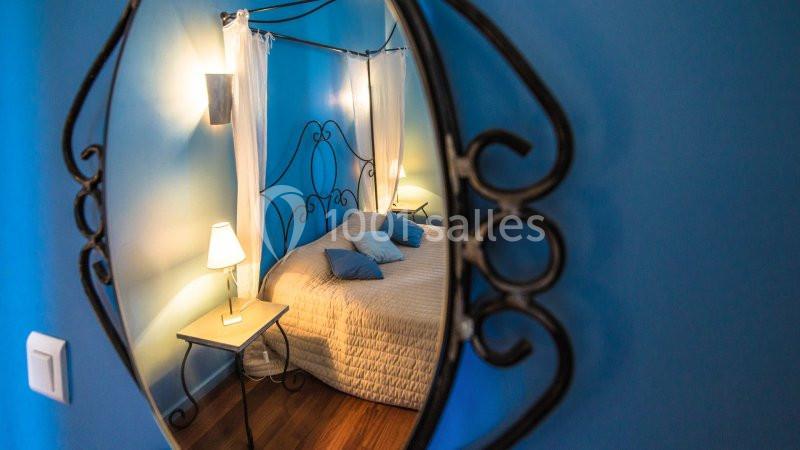 Reflet d'un lit à baldaquin avec draps blancs et coussins bleus dans un miroir encadré de métal noir, éclairé par des lampes.