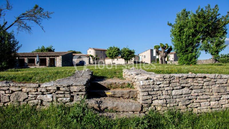 Mur en pierre sèche avec des marches, menant à un espace herbeux et des maisons en arrière-plan sous un ciel bleu.