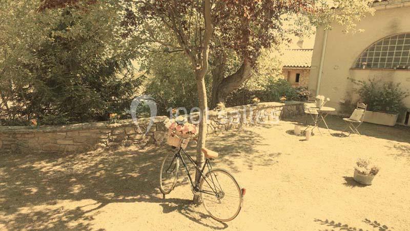 Un vélo avec un panier fleuri est posé sous un arbre dans une cour ensoleillée avec des chaises et une table en arrière-plan.