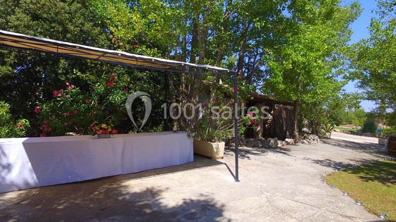 Table drapée de blanc sous une pergola, entourée de végétation et située dans une cour extérieure ensoleillée.