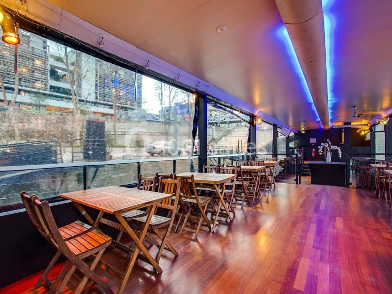 Salle de restaurant avec tables en bois, éclairage néon bleu et grandes baies vitrées donnant sur un extérieur urbain.