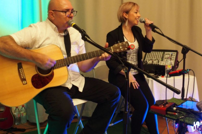 Miniature DJ Pézilla-la-Rivière (Pyrénées-Orientales) - Cleanjack #8 Un duo musical se produit sur une scène intérieure avec guitare et chant, devant un public assis.