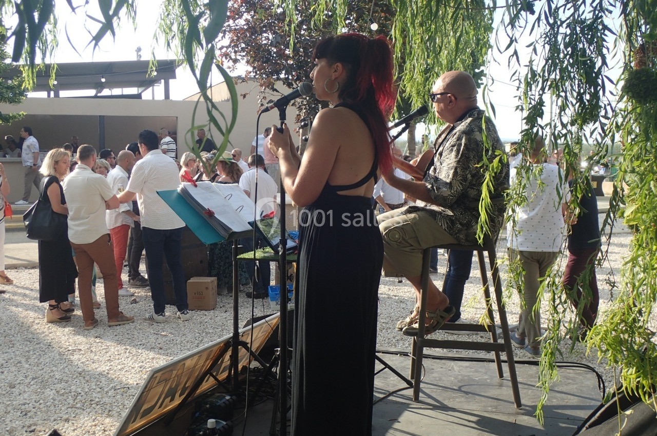 Une chanteuse et un musicien jouent sur une scène en plein air lors d'un événement avec des spectateurs en arrière-plan.